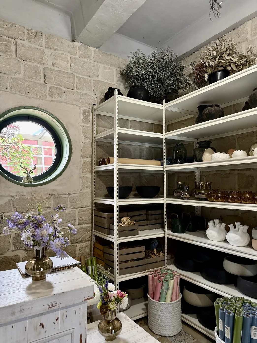 Rafturi BOX albe cu vaze și boluri lângă peretele de piatră al atelierului Florile Mele, lângă o fereastră rotundă