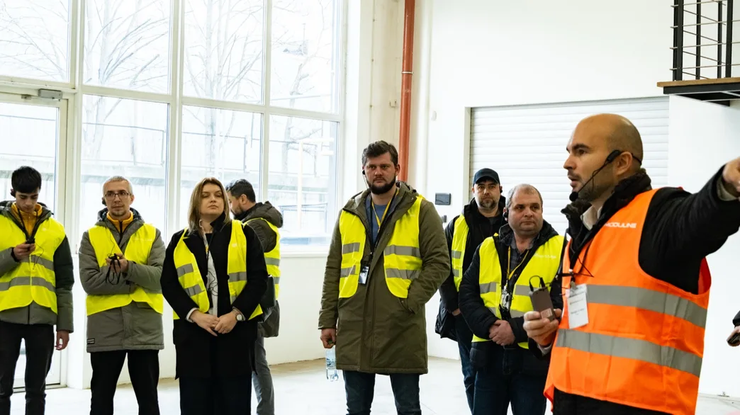 Vizitatori în veste reflectorizante ascultând un ghid Moduline în hala de producție